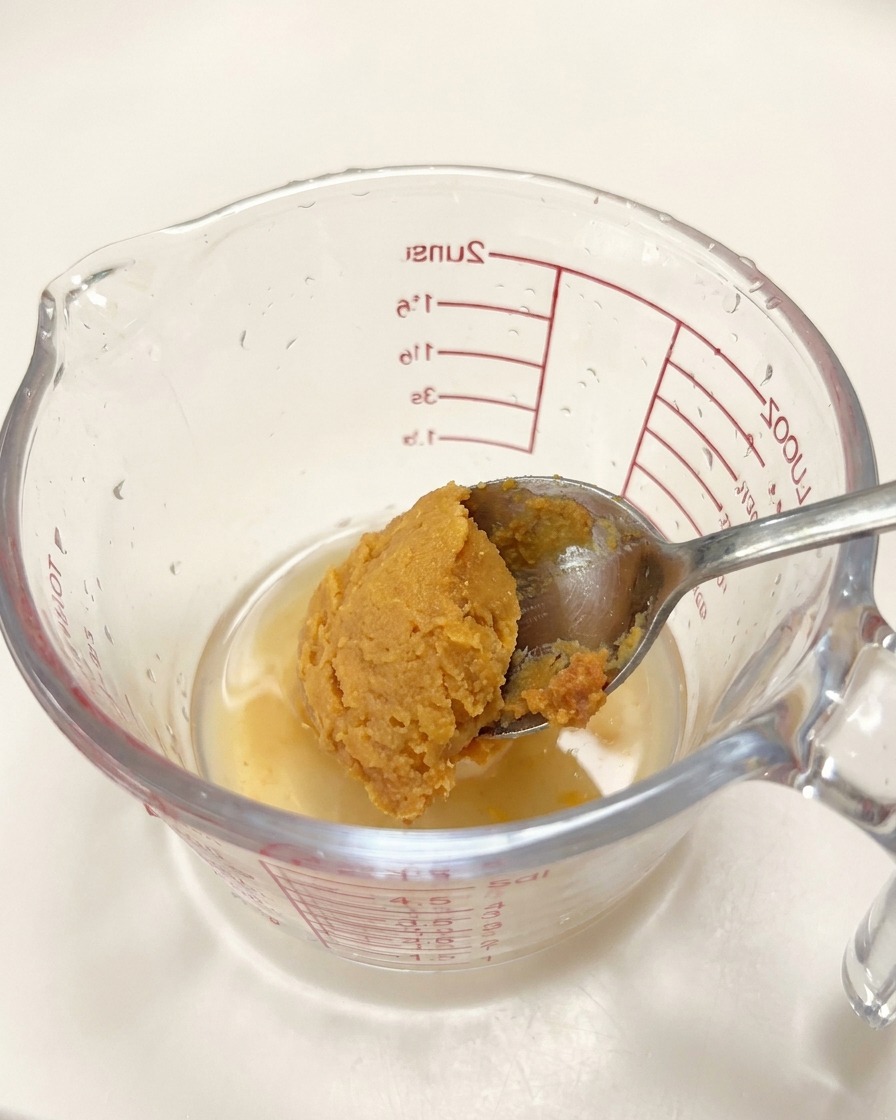 A spoonful of miso paste being added to a glass measuring cup containing liquid seasonings.