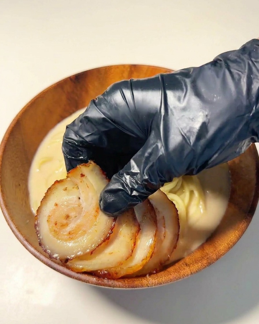 A black-gloved hand arranging slices of seared chashu pork on top of the ramen noodles.