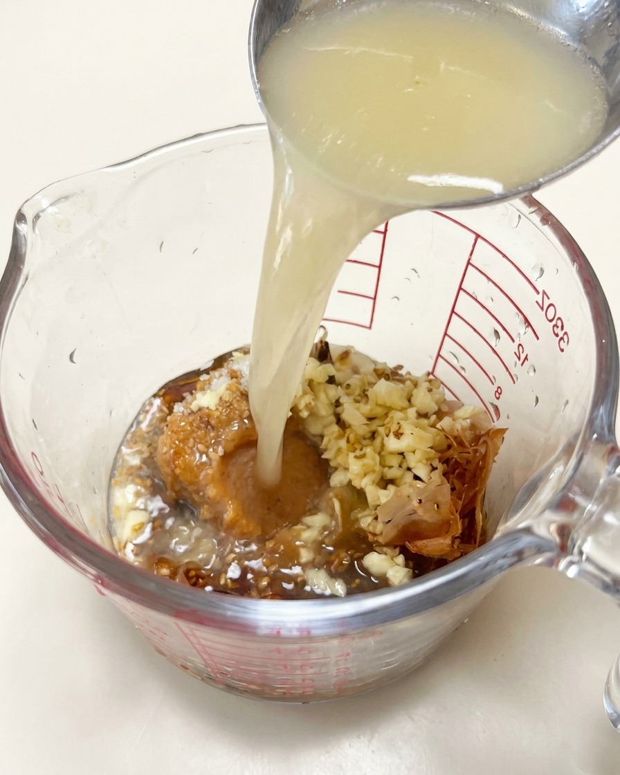 Creamy pork broth being ladled into a measuring cup containing miso, bonito flakes, and minced garlic.