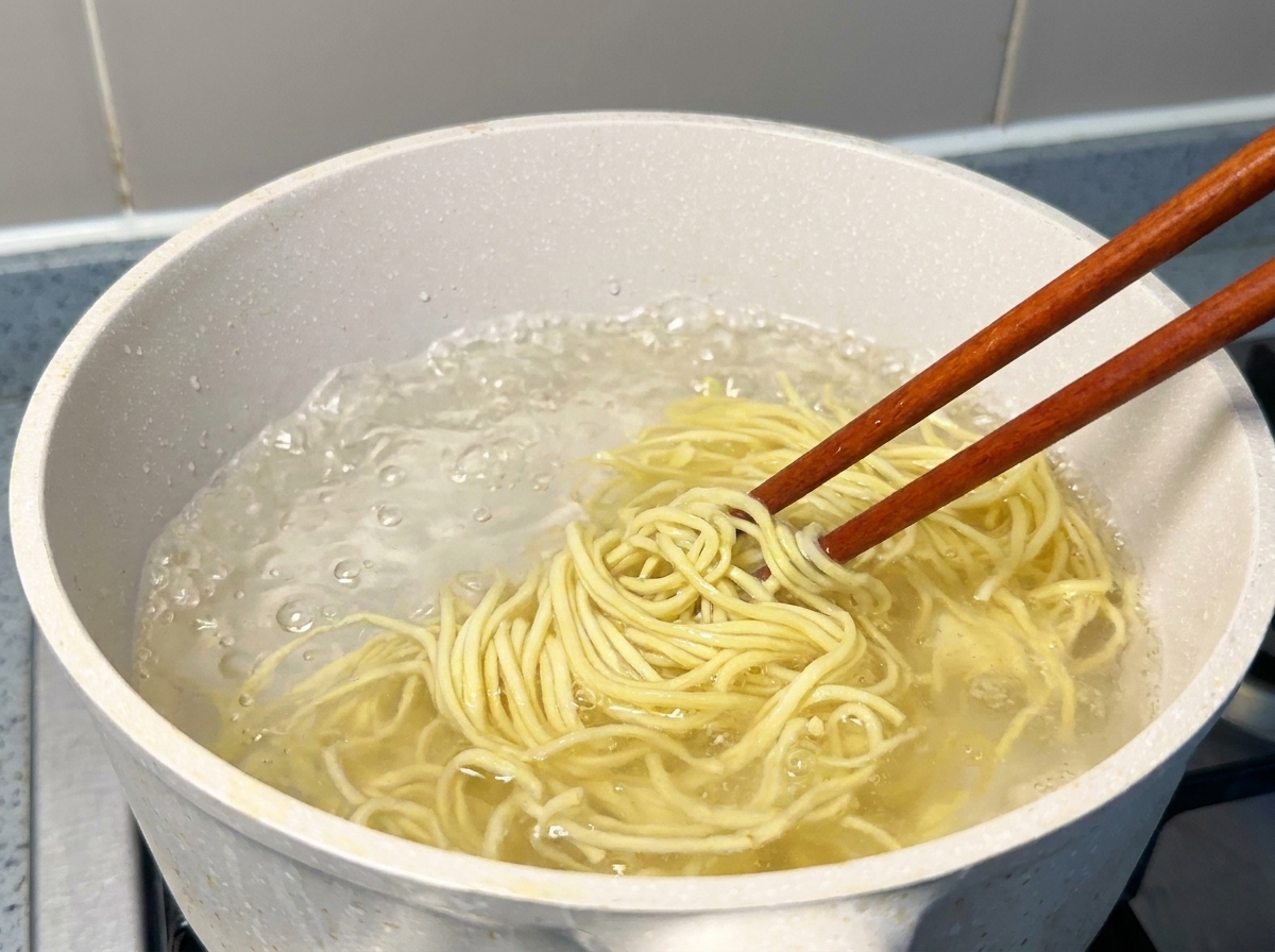 Fresh yellow ramen noodles being lifted with wooden chopsticks from a pot of vigorously boiling water.