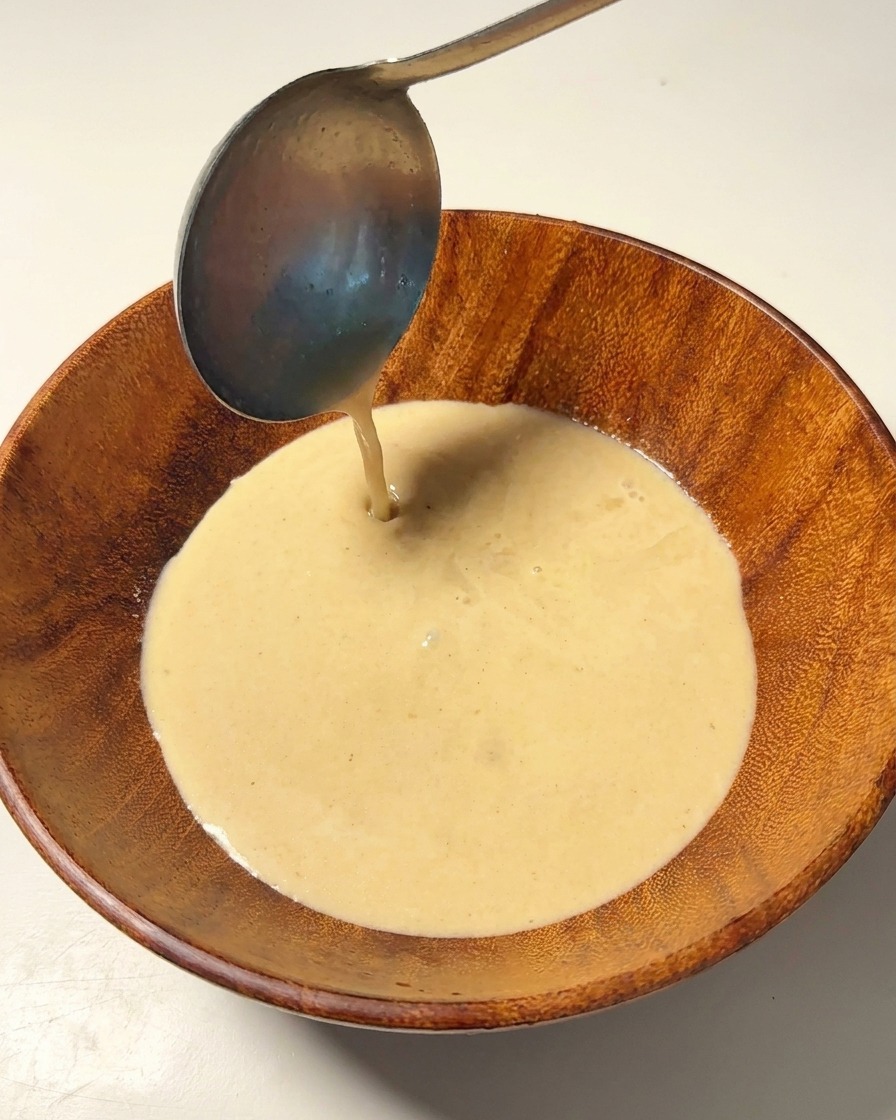 Hot, creamy pork bone broth being poured from a metal ladle into a wooden bowl.