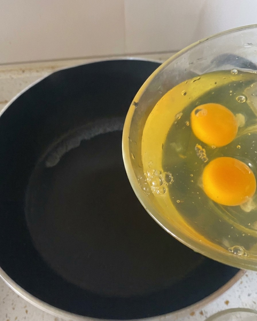 Two raw eggs being poured from a clear glass bowl into a hot, dark frying pan.