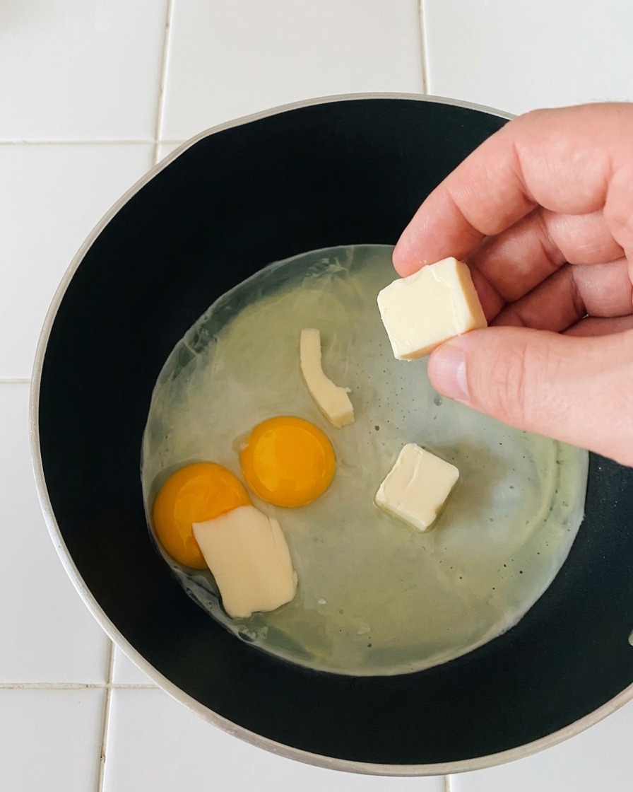 A hand dropping small, rectangular slices of white mozzarella cheese onto gently frying sunny-side-up eggs.