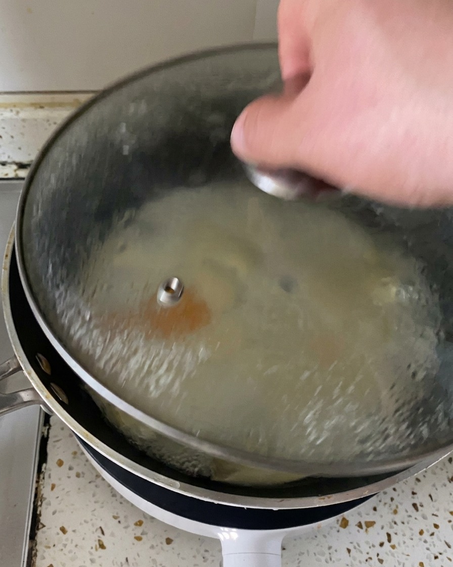A hand lifting a glass lid off a pan to reveal steam and perfectly cooked eggs topped with melted cheese.