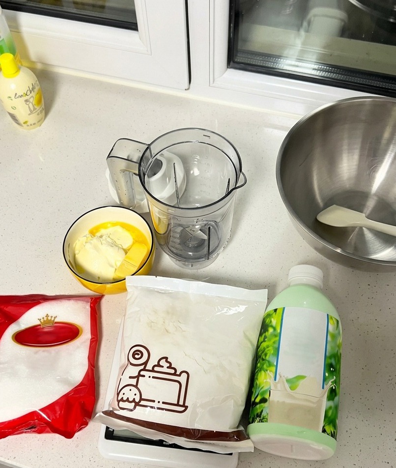 A countertop spread of baking ingredients including a milk bottle, a bag of sugar, flour, and a bowl with butter and cream cheese.