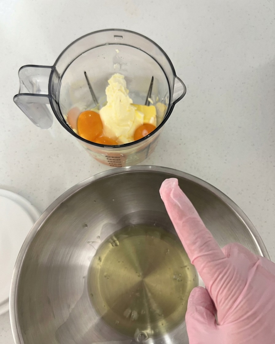 Egg yolks, butter, and cream cheese placed inside a clear blender cup, next to a stainless steel mixing bowl containing egg whites.