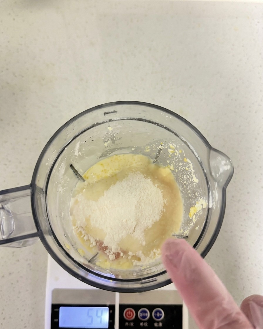 Flour and other dry ingredients sitting on top of the wet mixture inside a blender cup resting on a digital scale.