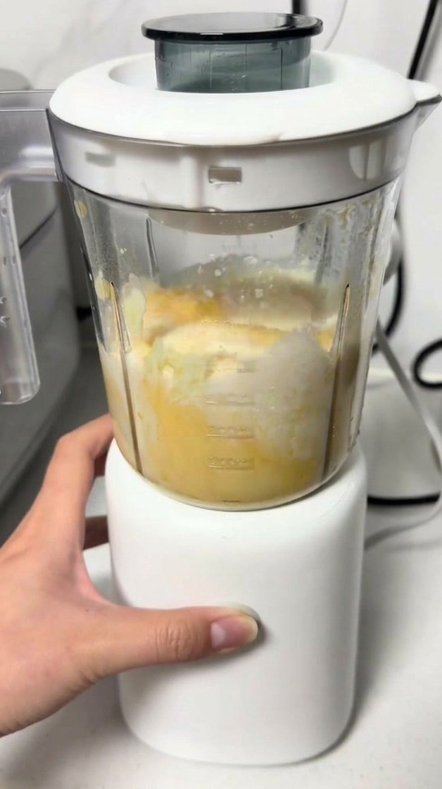 A hand operating a small white blender as it mixes a yellow cheese and egg batter inside the clear cup.