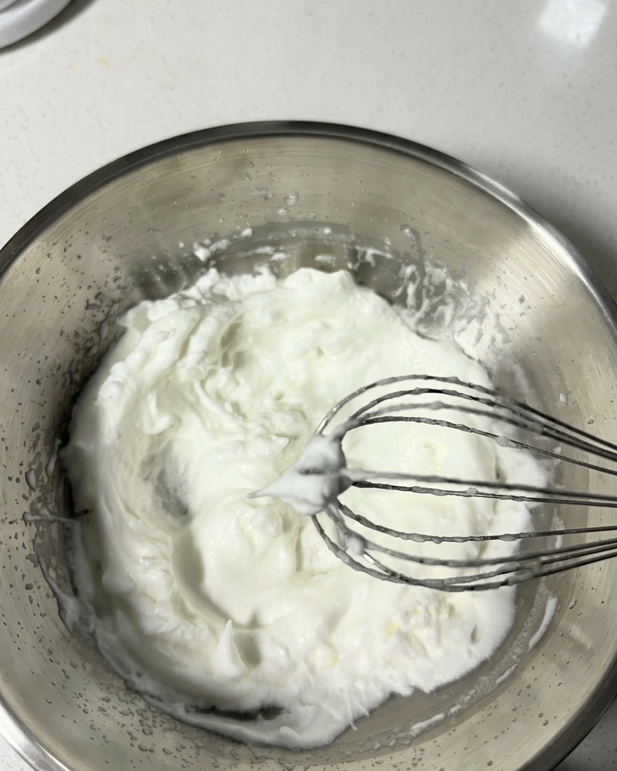 A metal whisk lifting whipped egg whites from a stainless steel bowl, displaying clear soft peaks.