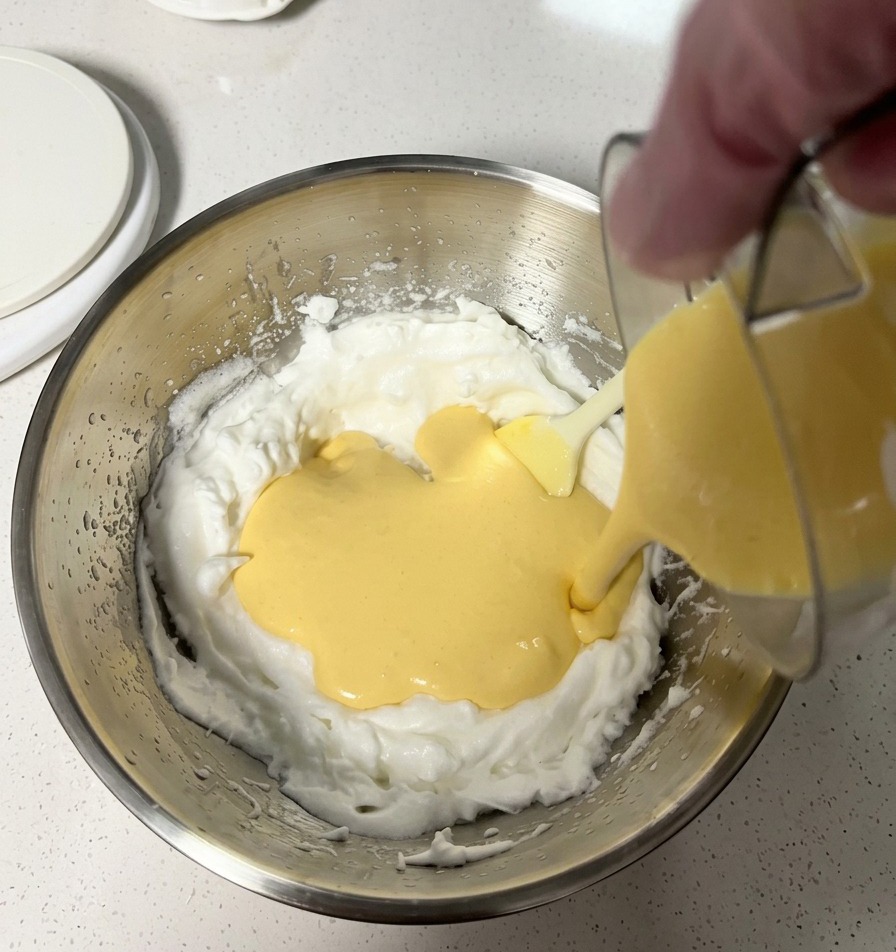 Smooth yellow batter being poured into a metal mixing bowl filled with fluffy white whipped egg whites.