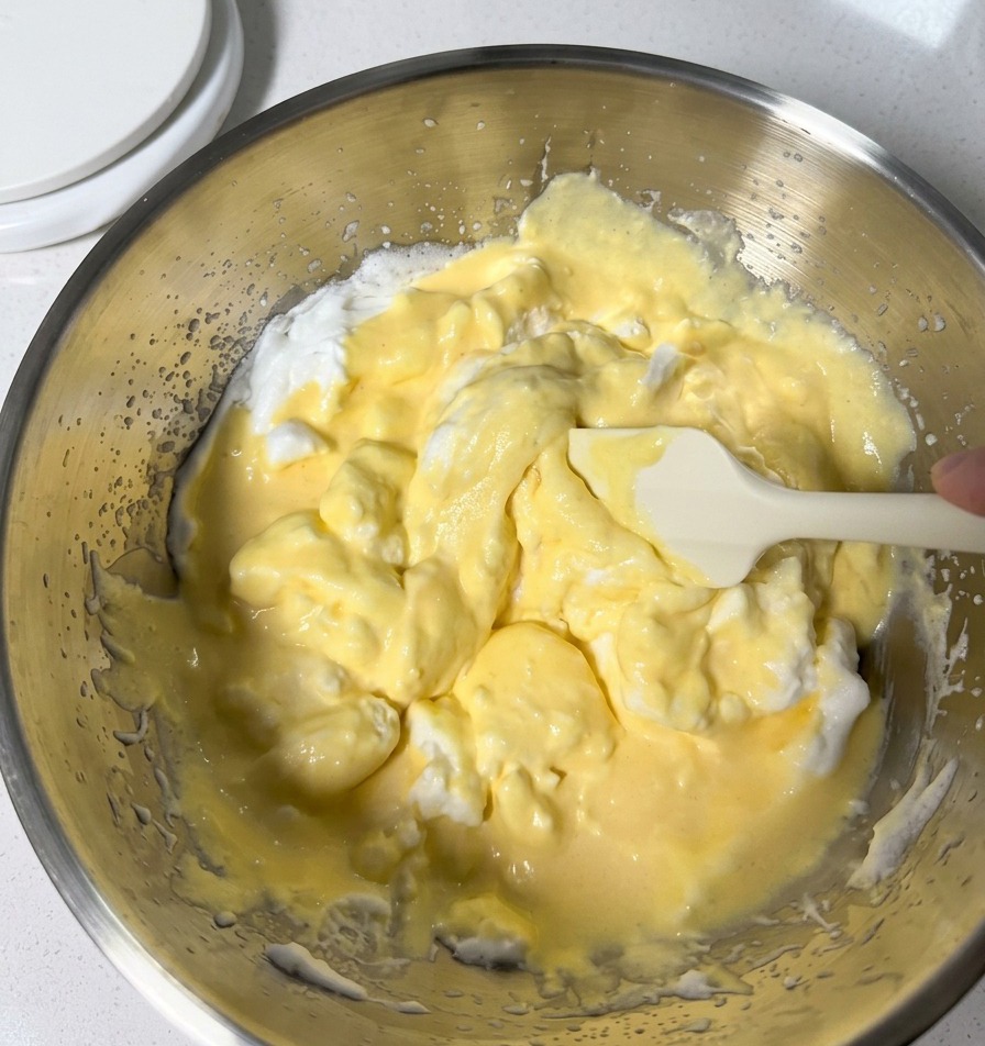 Gently folding pale yellow batter with fluffy white whipped egg whites in a metal mixing bowl using a white silicone spatula.