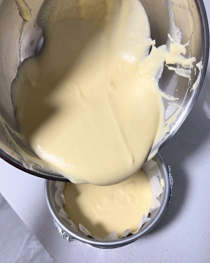 Smooth, pale-yellow cheesecake batter being poured from a stainless steel mixing bowl into a round 6-inch cake pan lined with crinkled baking paper.