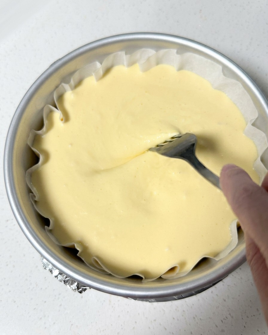 A hand using a metal fork to gently swirl the top surface of pale-yellow cheesecake batter inside a lined round cake pan.