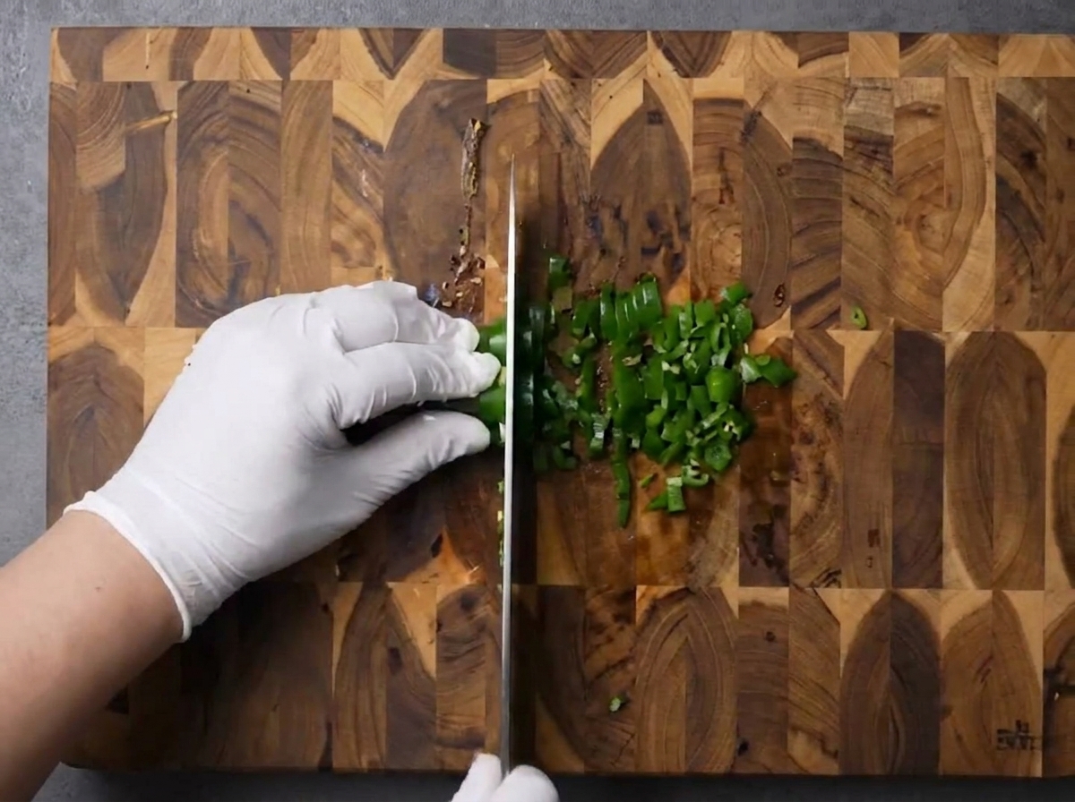 Gloved hands using a knife to finely chop green chilies on a patterned wooden cutting board.