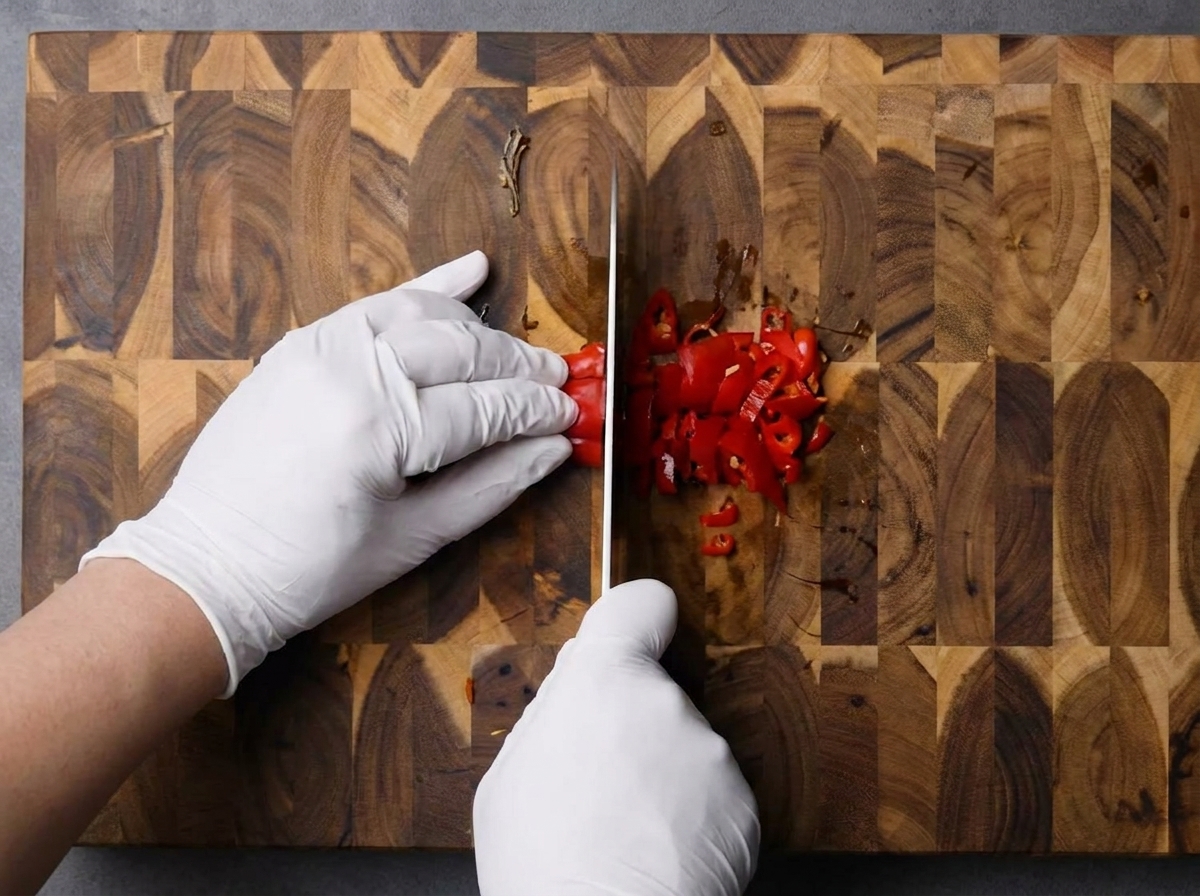 Gloved hands using a knife to finely chop fresh red chilies on a wooden cutting board.