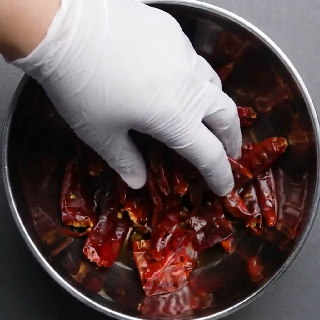 A gloved hand pushing whole dried red chilies down into a metal bowl of water to soak.