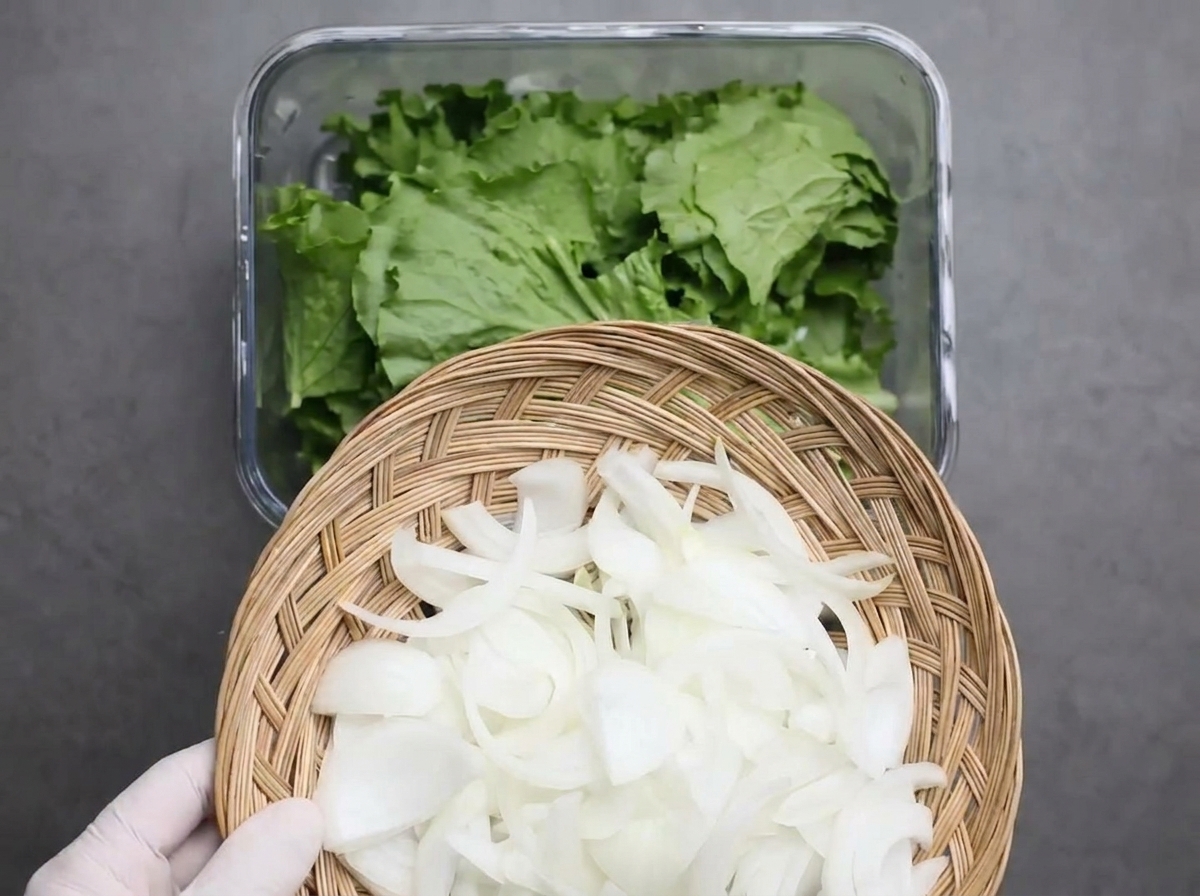 Sliced white onions being poured from a woven bamboo basket onto a bed of torn green lettuce leaves in a glass container.