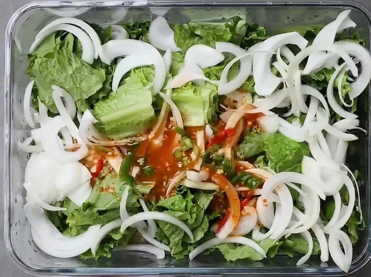 Red chili broth with chopped green chilies poured over fresh torn lettuce and sliced white onions in a rectangular glass food storage container.
