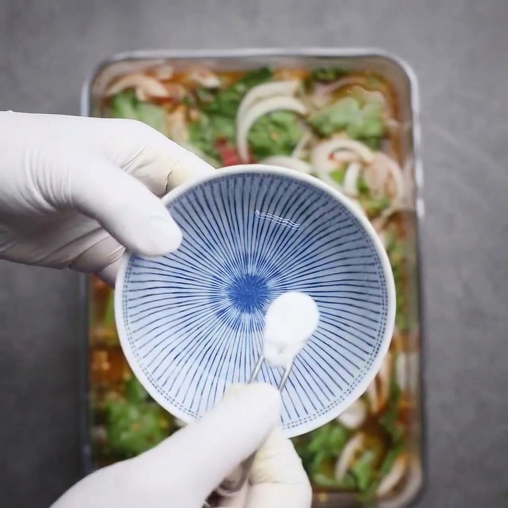 Gloved hands dissolving a small spoonful of white sweetener powder into a small blue and white patterned bowl filled with water.