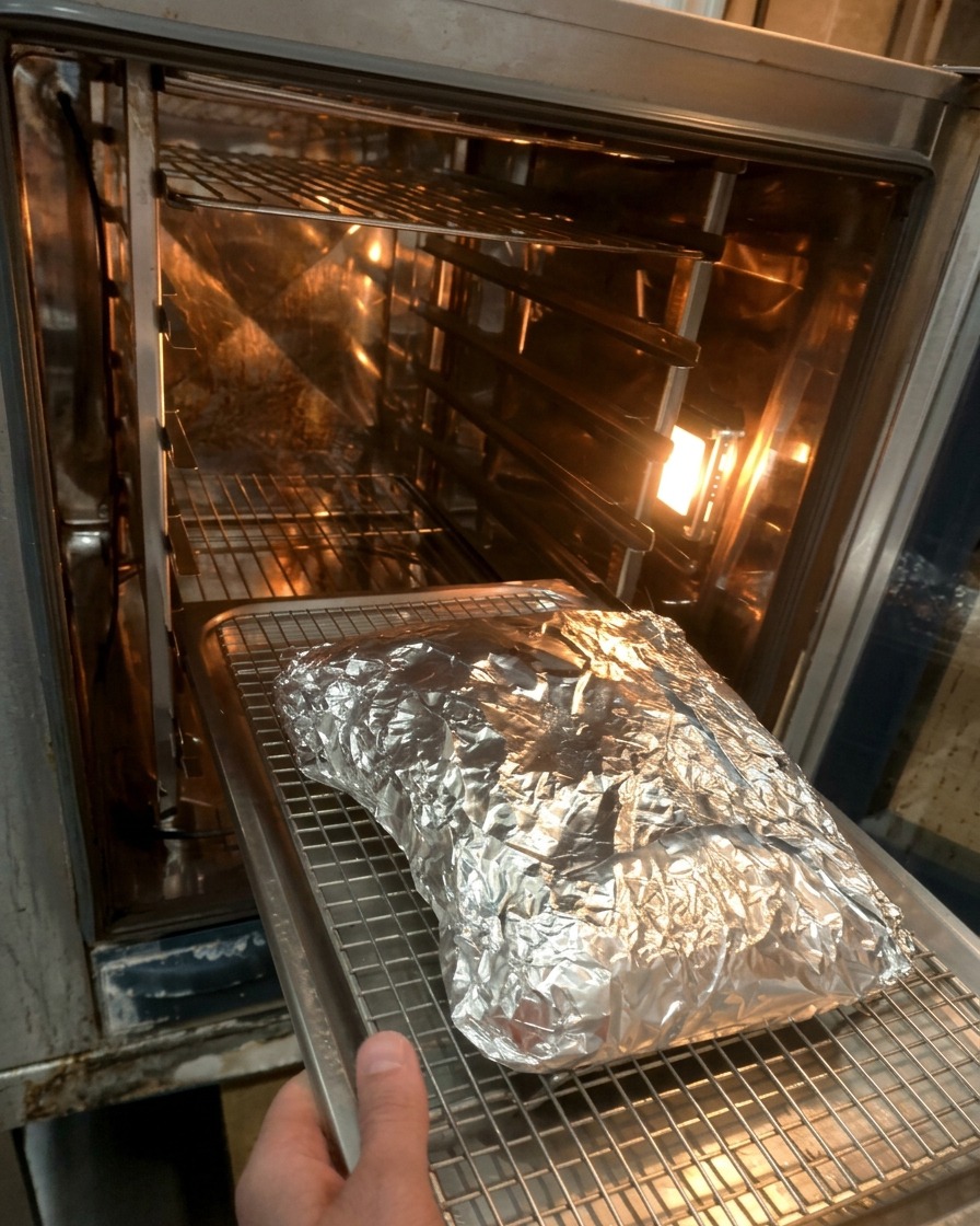 A foil-wrapped parcel of beef ribs being placed onto a wire rack inside a hot oven.
