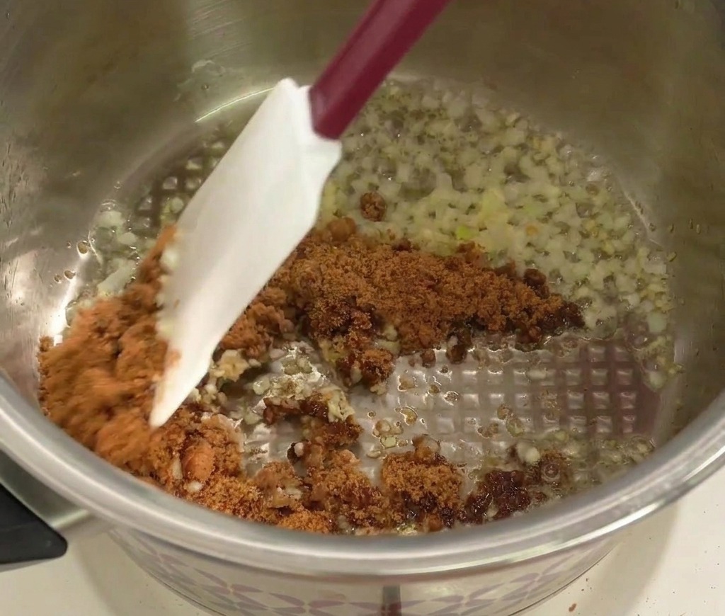 A spatula stirring dark brown sugar into sautéed minced onions and garlic in a stainless steel pot.