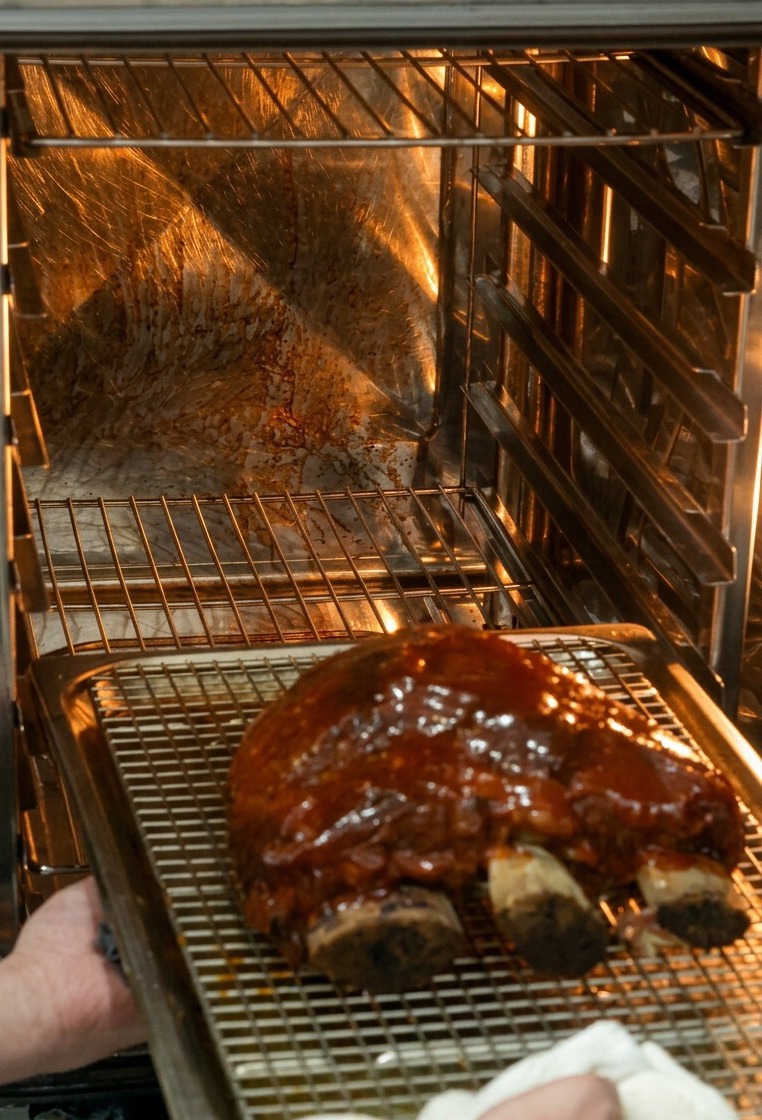 Hands sliding a baking tray with a wire rack holding a large, sauce-glazed beef short rib into a brightly lit hot oven.