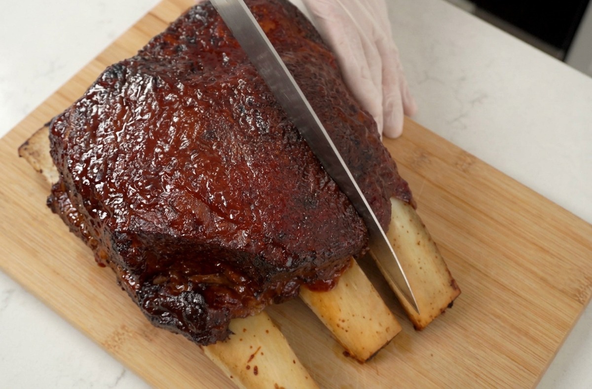 A gloved hand holding a sharp knife as it slices through a large, beautifully glazed rack of roasted beef short ribs on a wooden board.