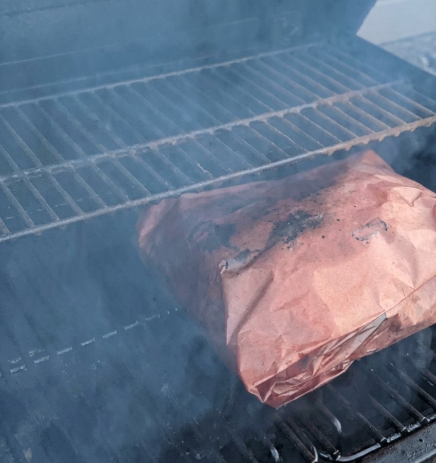 Butcher-paper-wrapped beef ribs cooking on smoker grates surrounded by smoke.