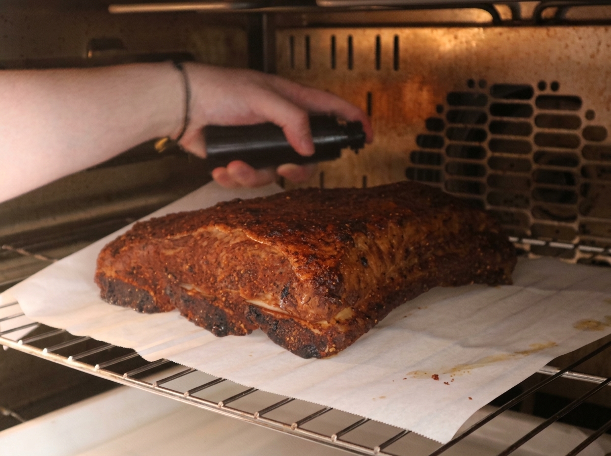 A hand holding a black spray bottle, spritzing a mixture onto a spice-coated beef rib roasting inside an oven.
