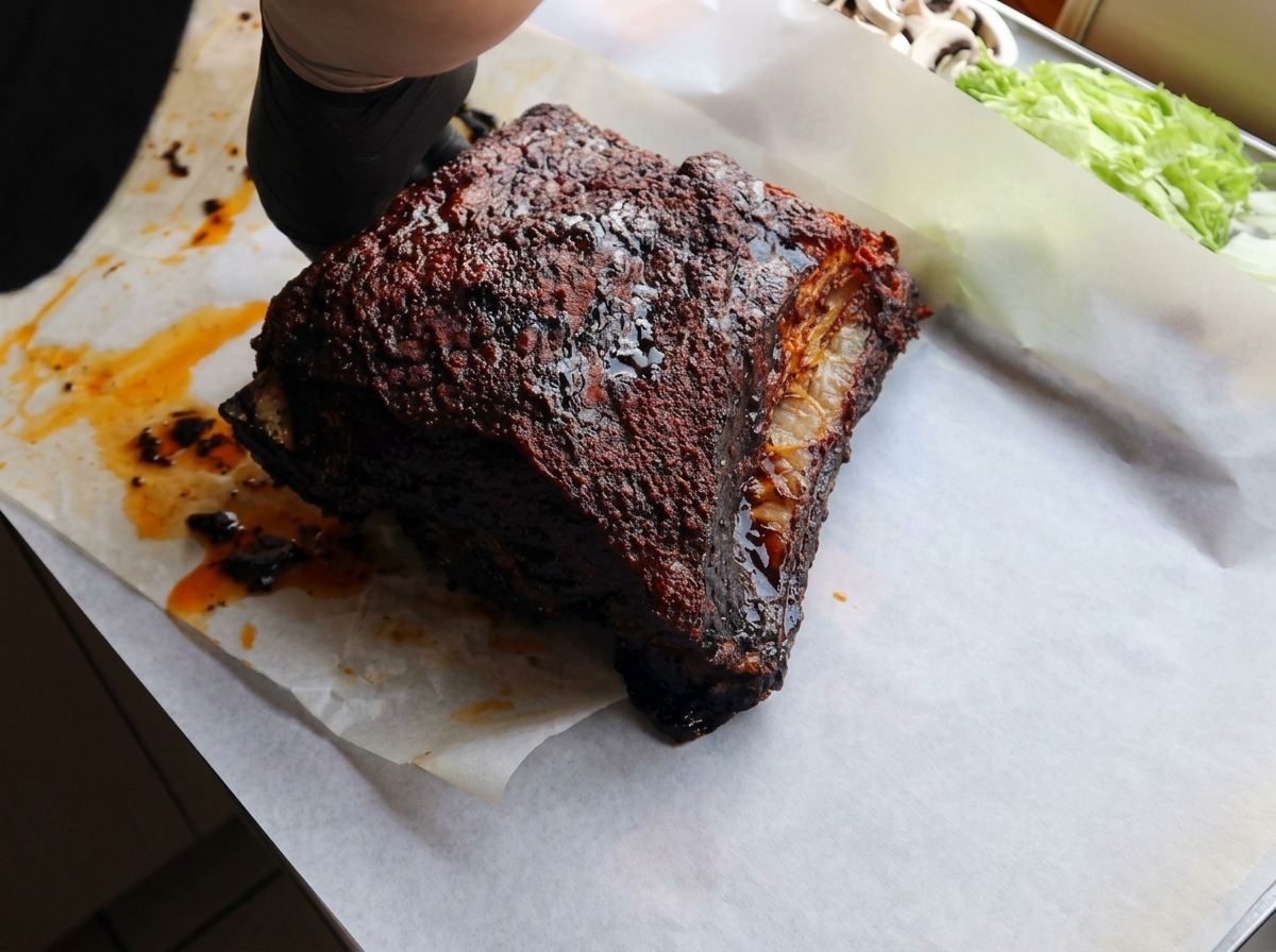 Hands wearing black gloves placing a dark, roasted beef rib onto a sheet of white parchment paper.