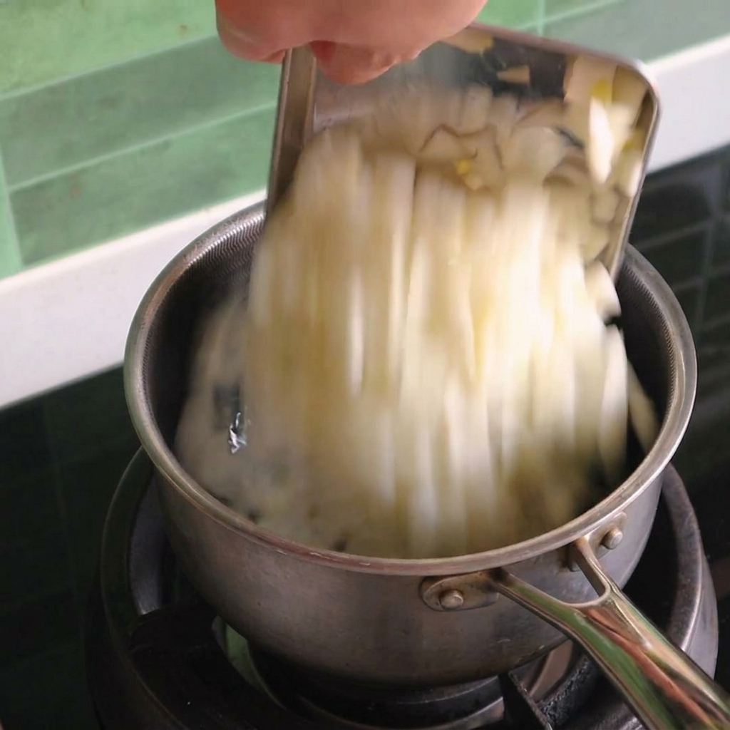 Diced white onions being poured from a metal scraper into a small stainless steel pot on a gas stove.