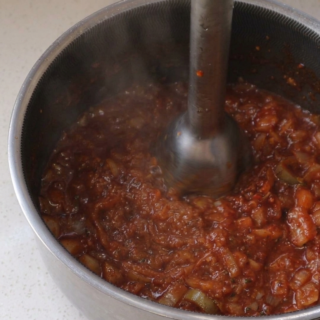 Using a metal immersion blender to puree a simmering, chunky red BBQ sauce inside a stainless steel pot.