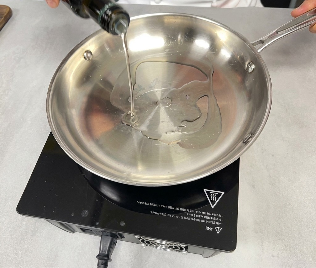 A hand pouring cooking oil from a dark green glass bottle into an empty stainless steel pan on a black induction stove.