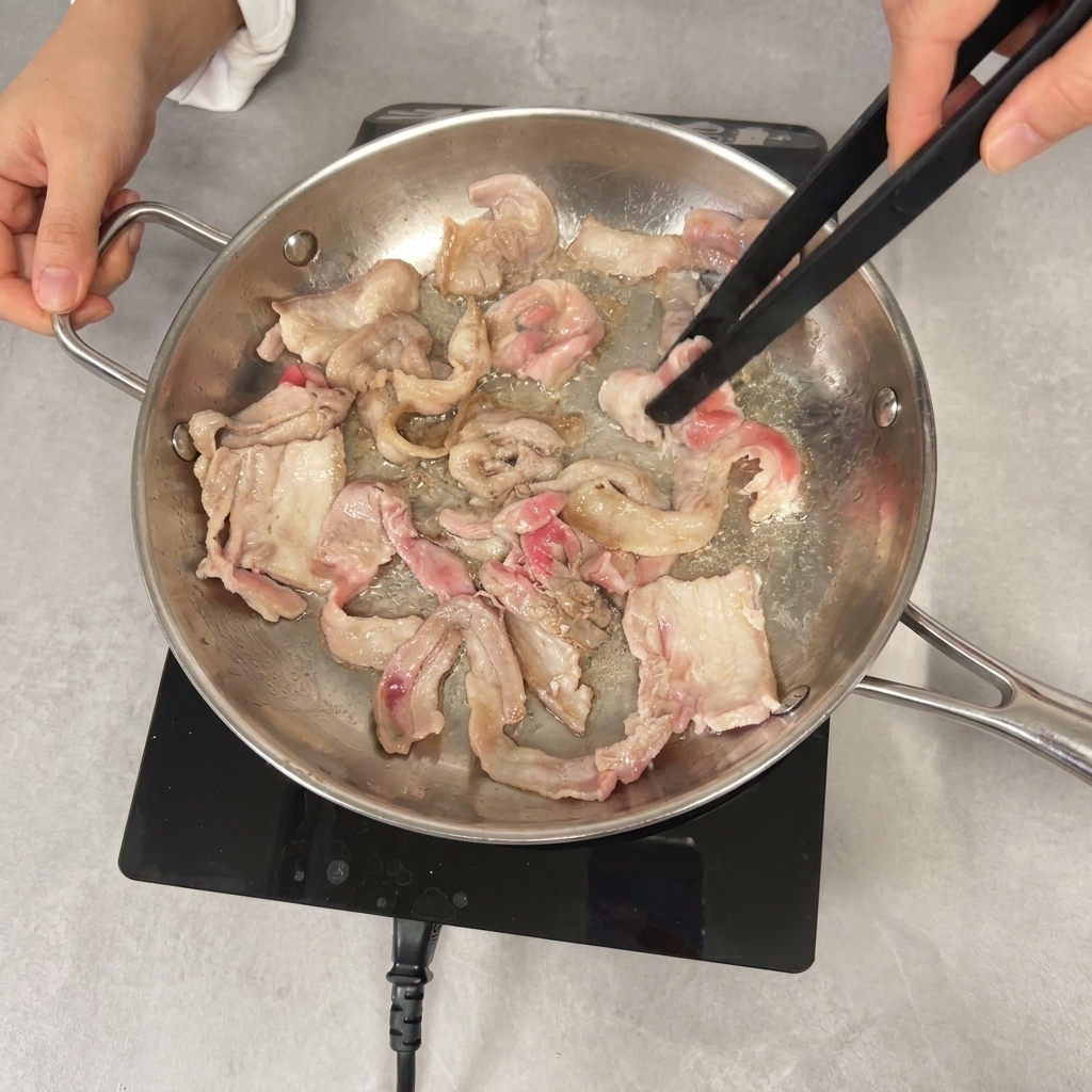 Using black tongs to stir-fry partially browned pork belly slices in a pan.