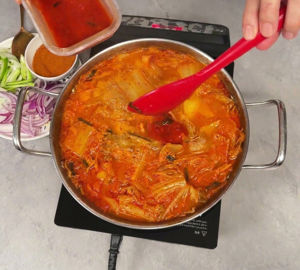 Bright red kimchi juice being poured from a small plastic container into a simmering pot of kimchi and vegetable stew, mixed with a red spatula.