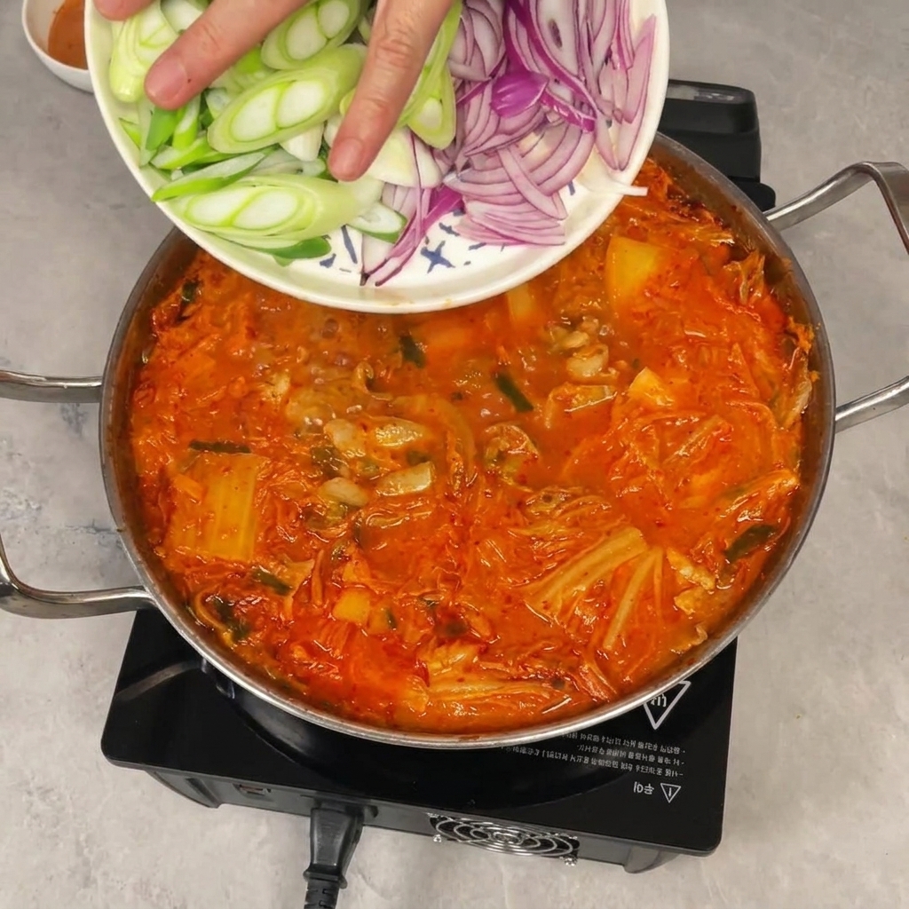 A hand sliding sliced green and red onions from a plate into a simmering pot of red kimchi stew.