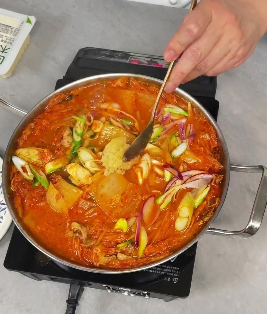Adding a spoonful of fresh minced garlic into a bubbling pan of red kimchi stew.