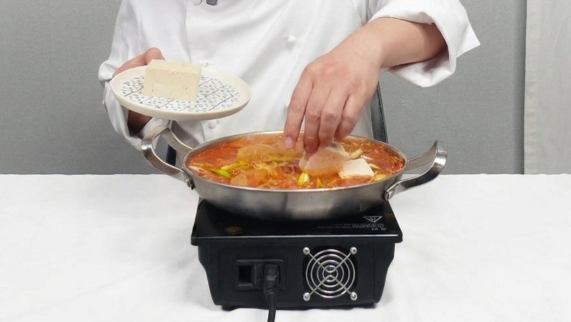Chef gently placing square slices of white tofu into a simmering pot of red kimchi stew.