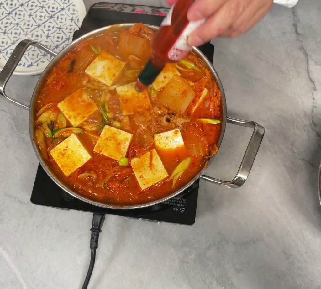A person dashing Tabasco hot pepper sauce from a small bottle into a boiling pot of red kimchi stew topped with square pieces of white tofu.