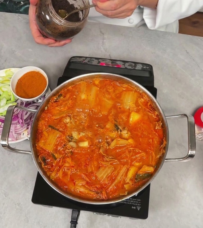 Chef holding a jar and scooping out a spoonful of thick dark soybean paste to add to the kimchi stew.