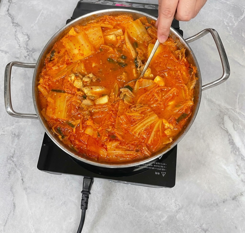 A hand using a stainless steel spoon to gently stir a vibrant red kimchi stew, mixing the seasonings into the broth.