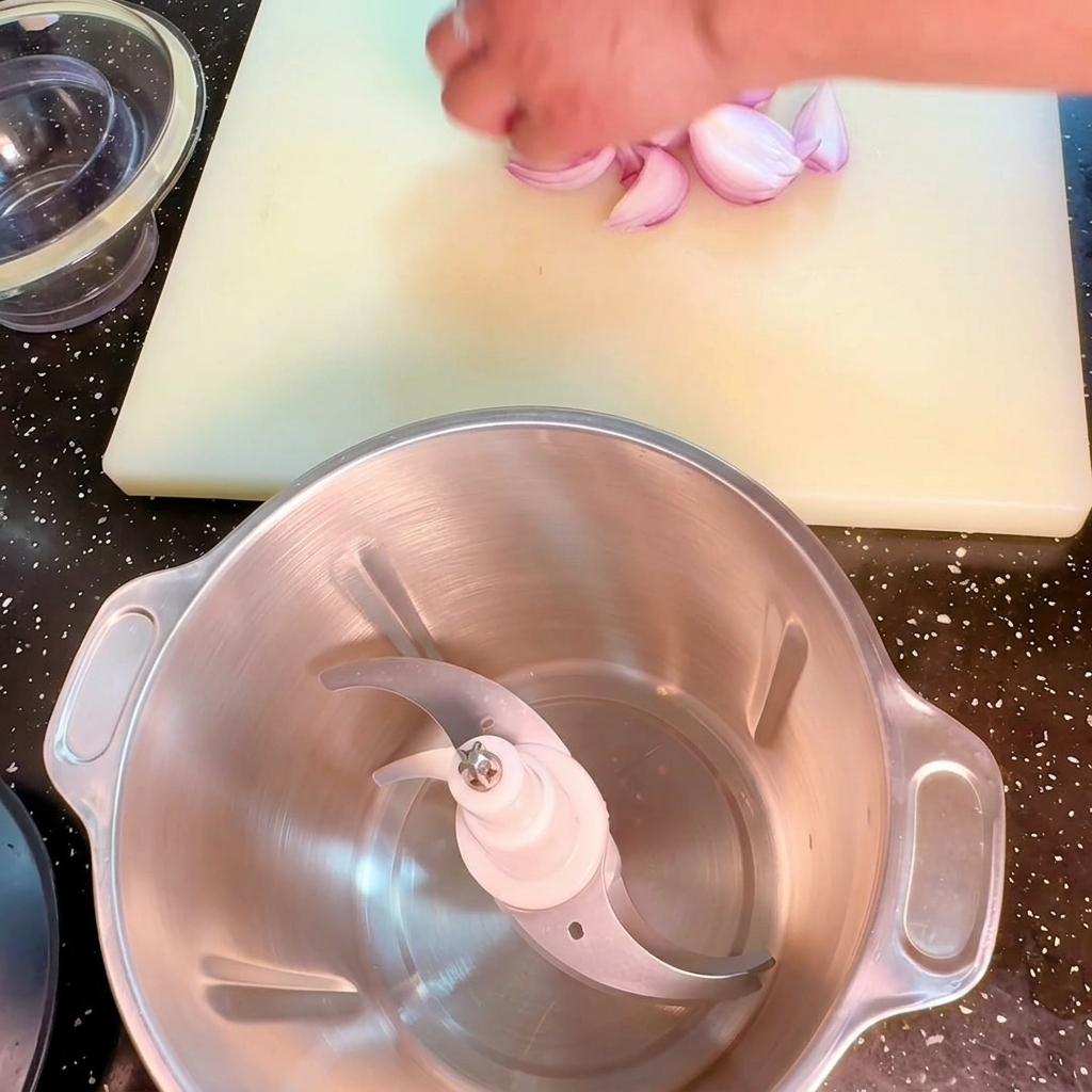 A hand placing chopped red onion pieces next to a stainless steel food processor bowl on a white cutting board.