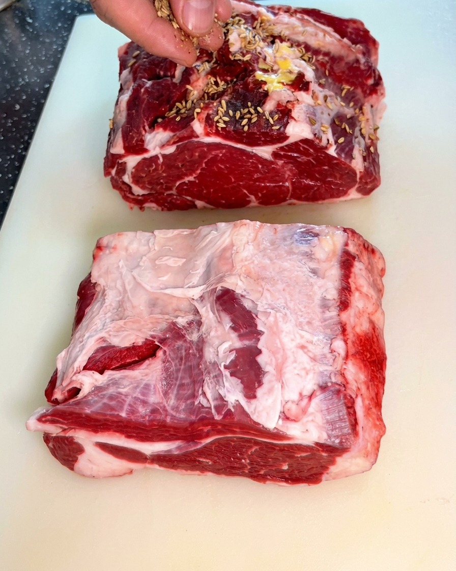 A hand sprinkling whole fennel seeds over two large, thick pieces of raw beef chuck.