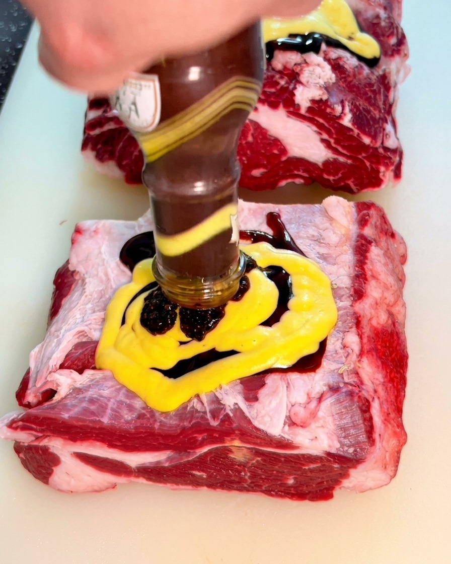 Thick black pepper sauce being poured from a glass bottle onto the yellow mustard layer on the beef.