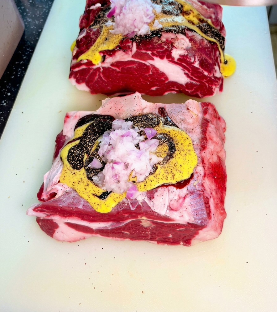 Raw beef topped with mustard sauce, dark soy, and minced red onions on a cutting board.