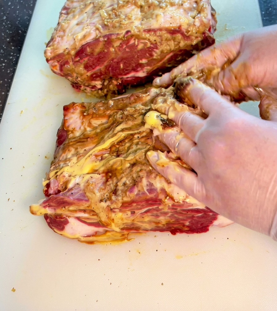 Hands actively rubbing a mixture of sauces and spices into a thick piece of raw beef on a white cutting board.