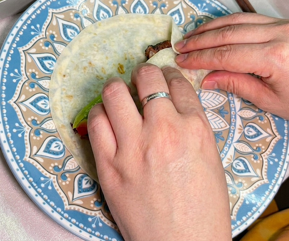 Two hands carefully rolling a tortilla filled with smoked beef and fresh vegetables into a tight wrap.