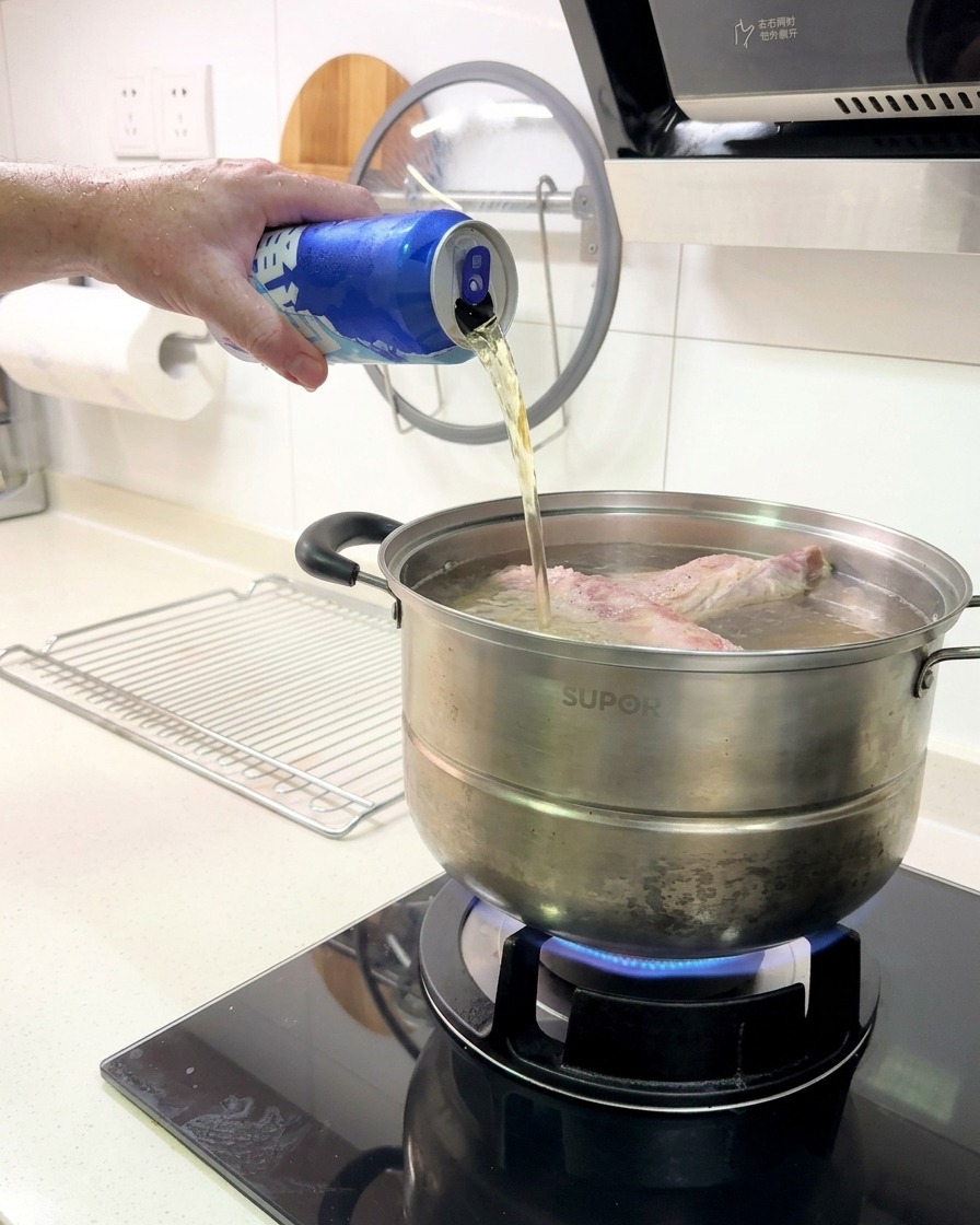 Beer being poured from a blue aluminum can into a pot containing a rack of ribs boiling on a gas stove.