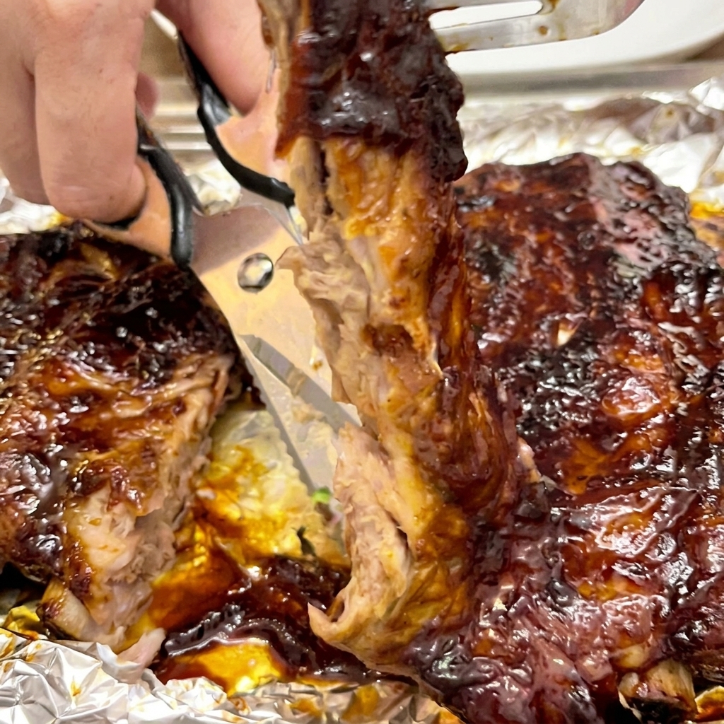 Close-up of kitchen scissors cutting through a tender, sauce-covered rib to separate it from the rack.