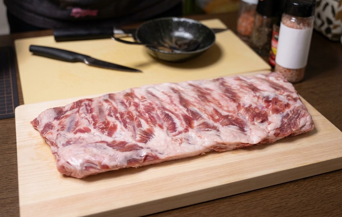 A whole rack of raw pork ribs laid flat on a wooden cutting board with a knife nearby.